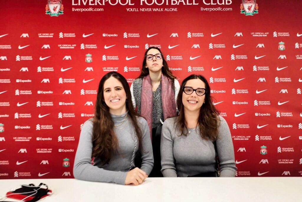 Anfield Stadium Tour - Press Room Three women in the Anfield press conference room against a red Liverpool FC sponsor backdrop