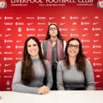 Anfield Stadium Tour - Press Room Three women in the Anfield press conference room against a red Liverpool FC sponsor backdrop