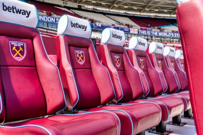 Premier League dugout with padded seats overlooking London Stadium pitch and home supporters section